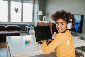 Young African american girl at home sitting on the table, using laptop, studying and looking at camera